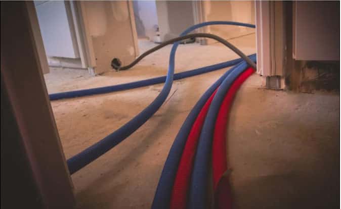 Gaines flexibles bleues et rouges (chauffage/plomberie) et un câble noir traversant un sol de chantier brut.