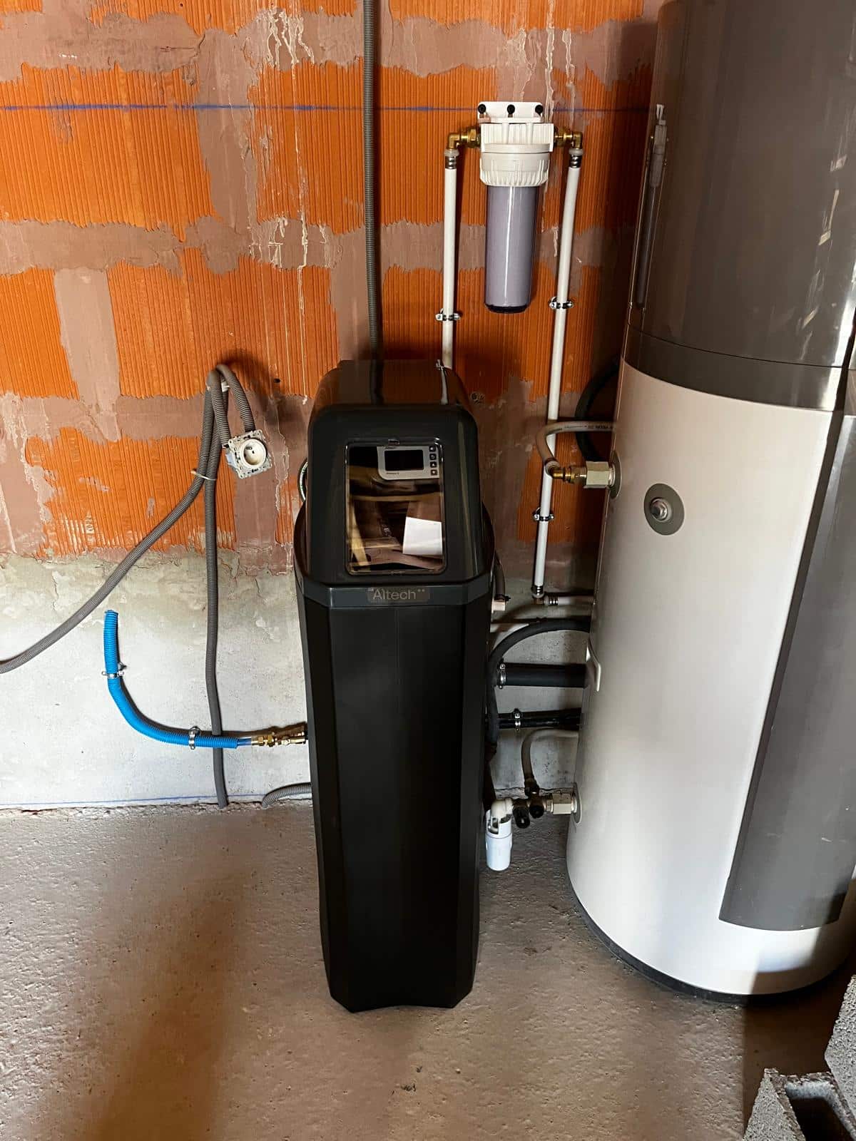 Adoucisseur d'eau noir Altech installé près d'un ballon d'eau chaude, avec filtre supérieur et tuyauterie visible sur mur de briques.