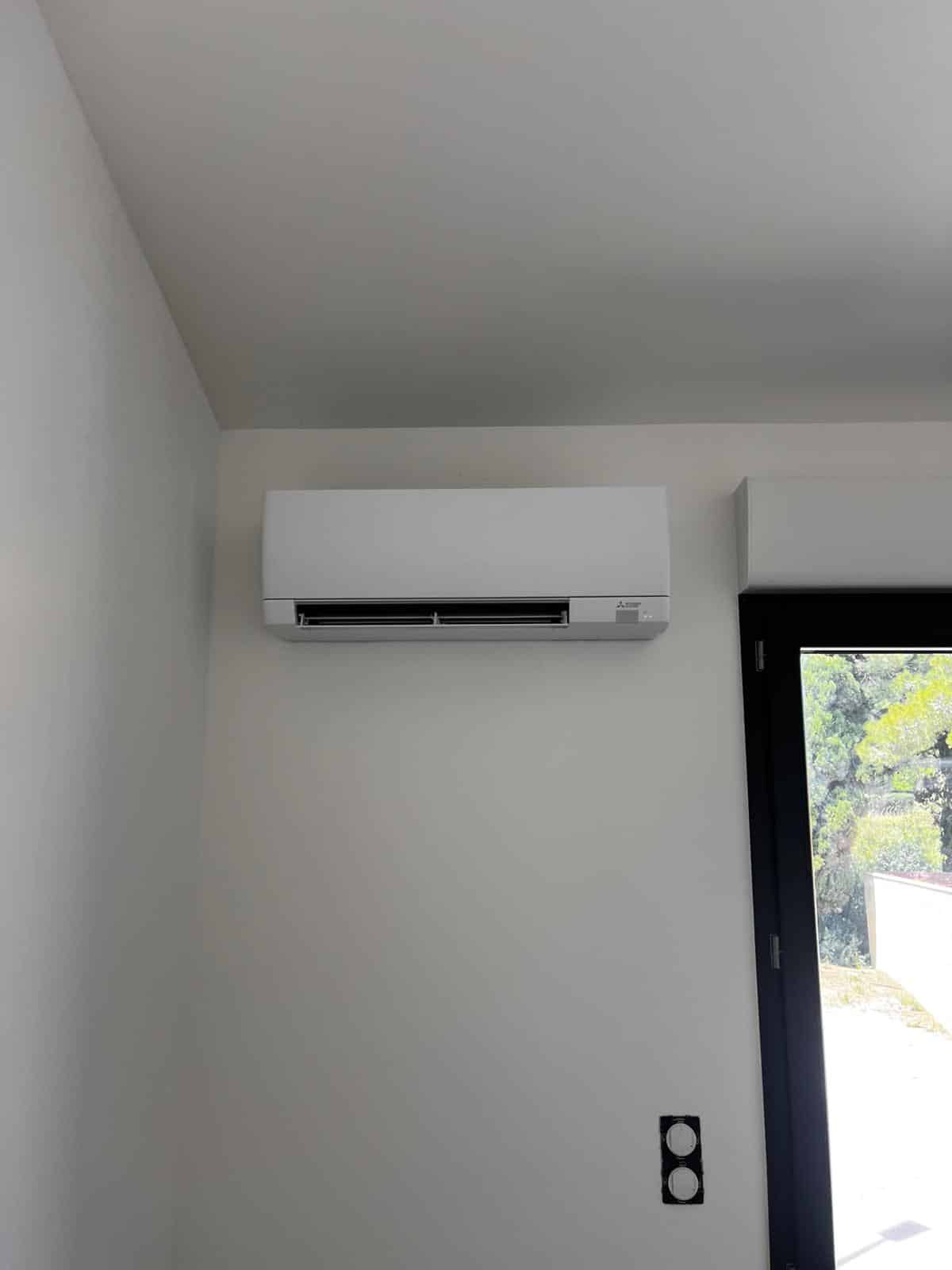 Climatiseur mural Mitsubishi blanc installé au-dessus d'une prise électrique noire et à côté d'une porte-fenêtre noire.