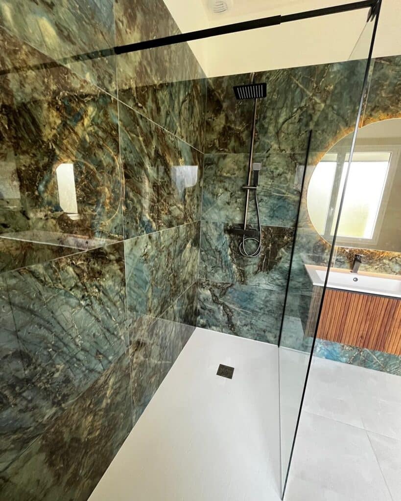 Salle de bain luxe : douche marbre vert et bois moderne Douche moderne avec parois vitrées noires et magnifique carrelage mural effet marbre vert-bleu profond, receveur blanc et meuble vasque bois.