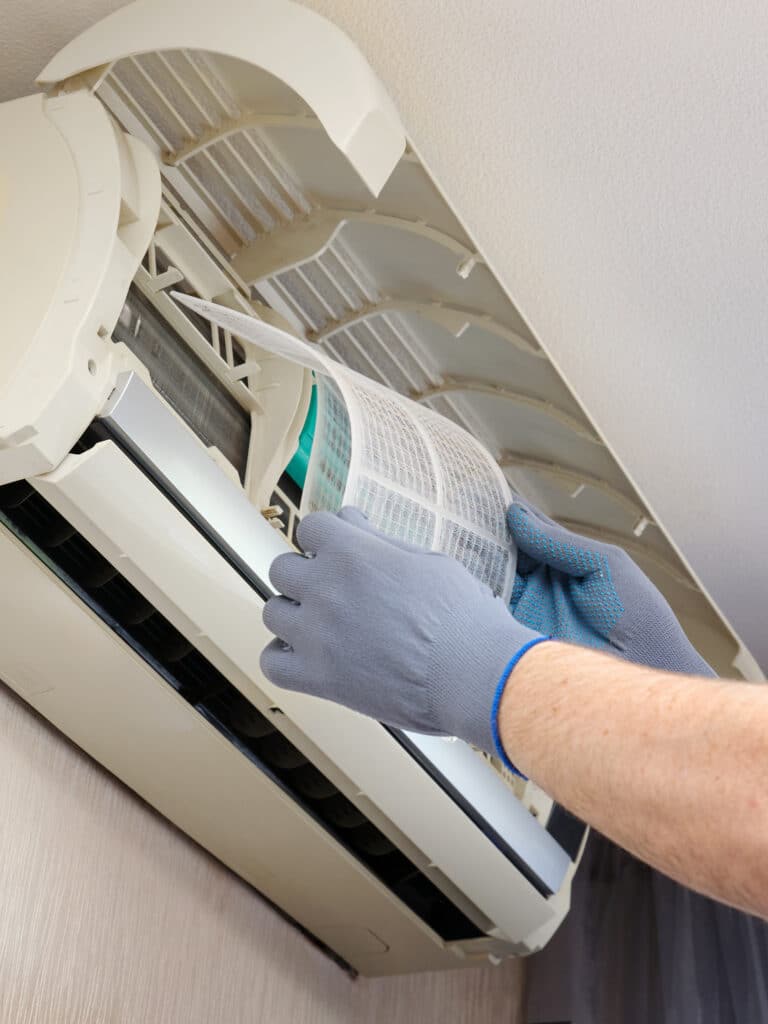 Nettoyage filtre climatiseur : entretien essentiel à domicile Mains gantées retirant un filtre à air sale d'une unité intérieure de climatiseur mural pour nettoyage.