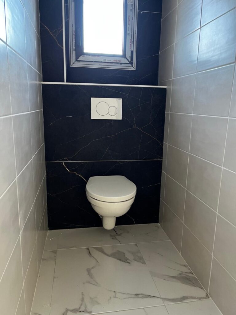 WC Moderne: Toilette Suspendue et Carrelage Noir/Gris Toilette suspendue dans un WC moderne, murs carrelés gris et panneau mural noir marbré avec boutons de chasse d'eau Geberit.